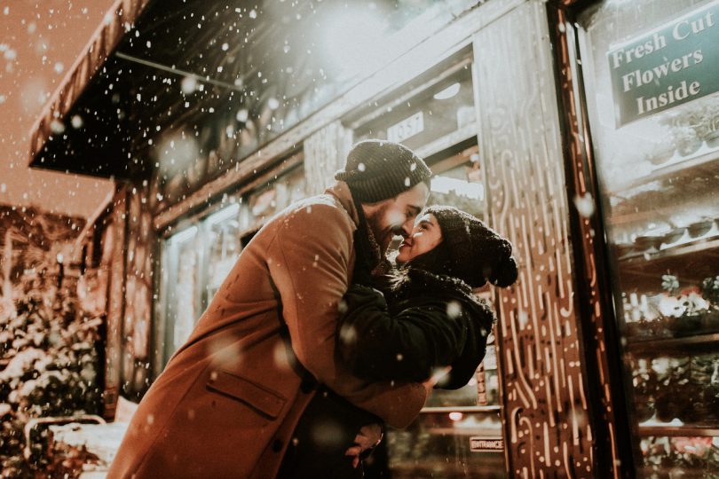 Casal se beijando com a neve caindo em cima deles.