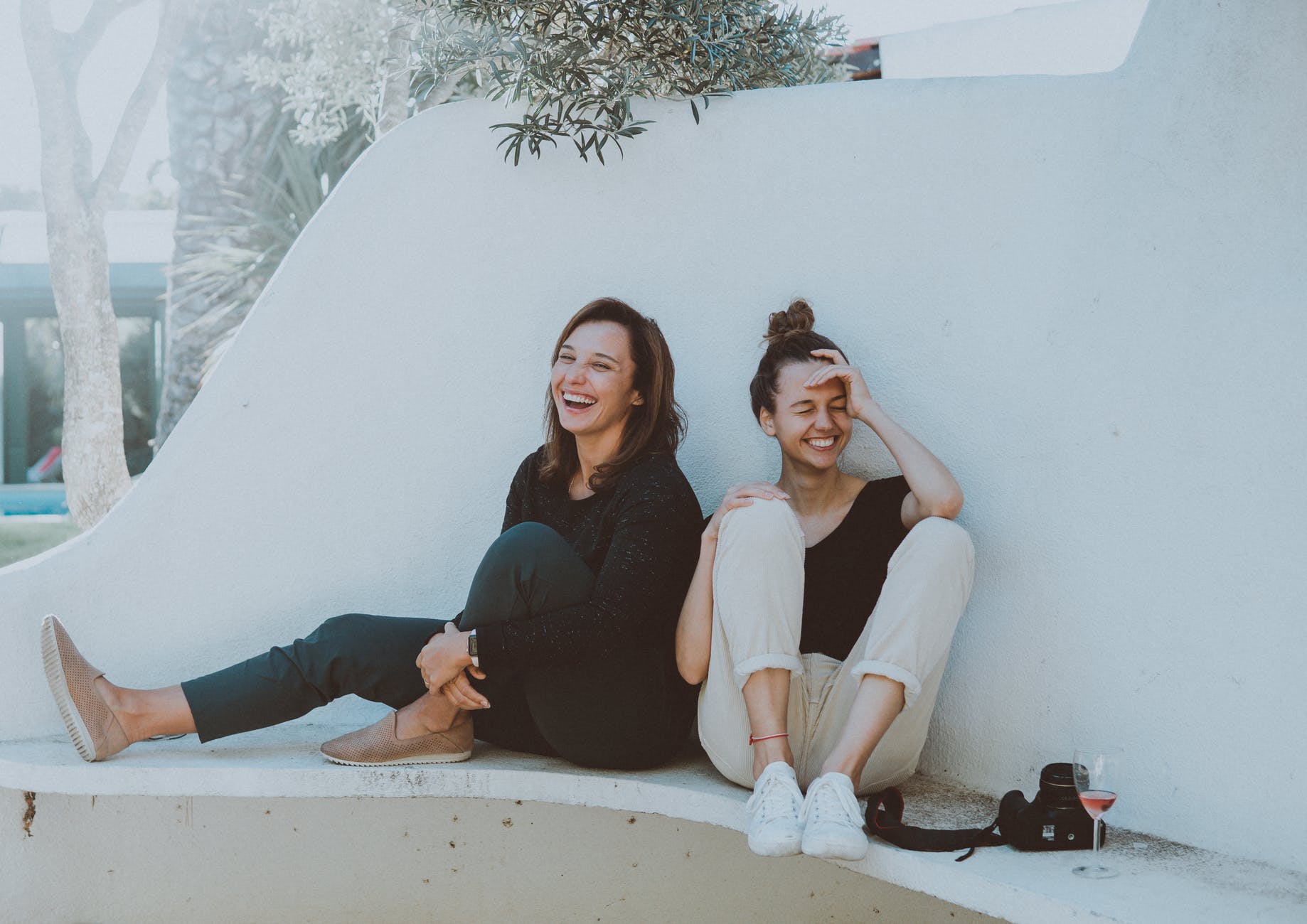 Duas mulheres brancas, jovens, ambas rindo, sentadas a bera de uma piscina vazia.