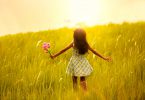 Garota pequena com os cabelos escuros e um veztido zadrez de cor branca e verde de costas correndo sobre as gramas sentido o pôr do sol,.