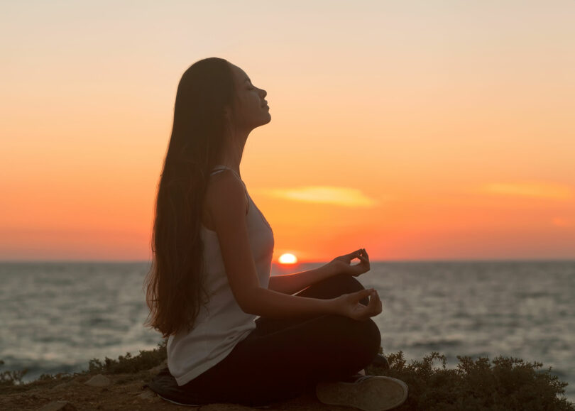 Mulher meditando sob montanha com mar e sol se pondo ao fundo