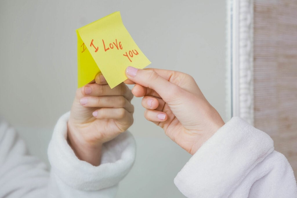 Mão feminina arrancando post it de espelho, que contém o escrito eu te amo, em inglês.