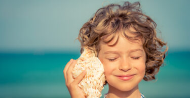 Menina sorrindo e ouvindo concha do mar na praia.