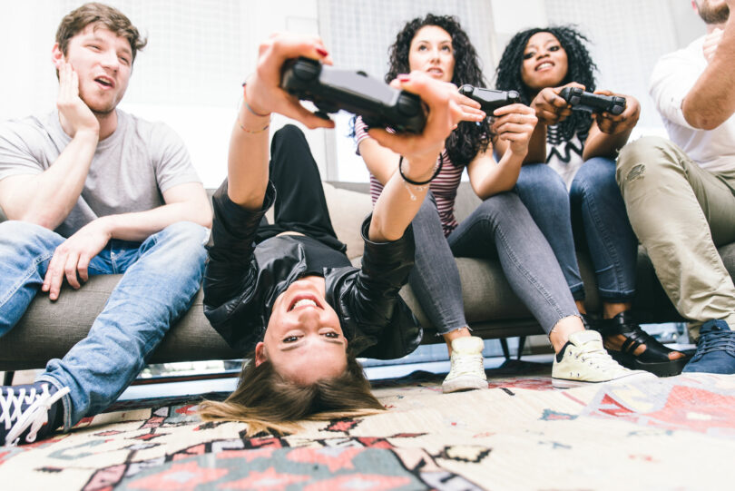 Quatro amigos jovens sentados em um sofá, três deles estão segurando um controle de videogame.