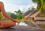 Mulher sentada praticando yoga em frente a duas casas cercadas pela natureza e por uma piscina.