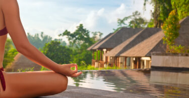 Mulher sentada praticando yoga em frente a duas casas cercadas pela natureza e por uma piscina.