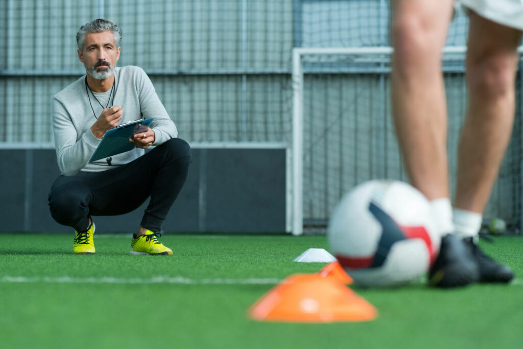 Homem, técnico de futebol, agachado no campo, segurando uma prancheta enquanto observa outro homem jogar