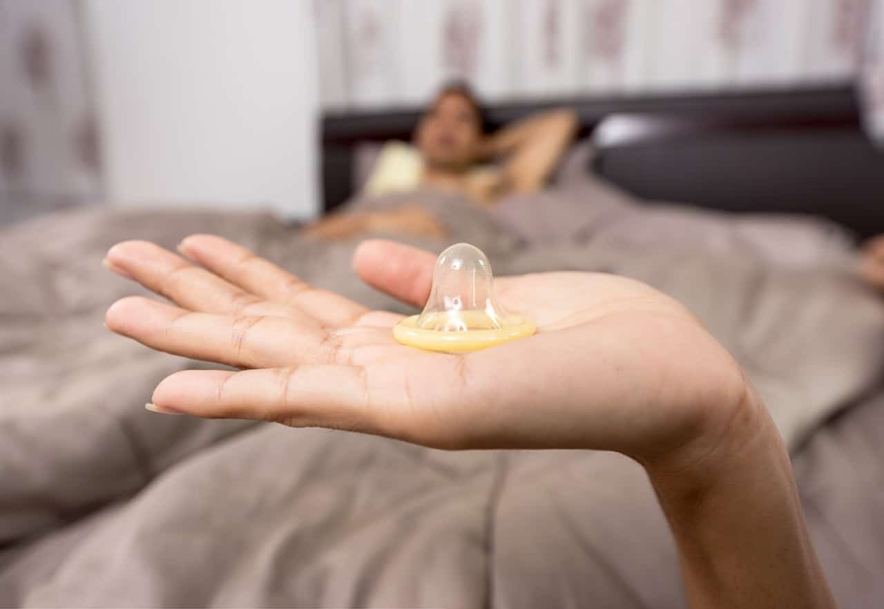 Mão segurando preservativo com homem deitado em cama ao fundo