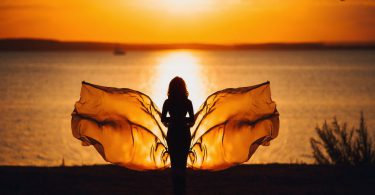 Mulher com "asas" de borboleta em frente ao mar no pôr-do-sol.