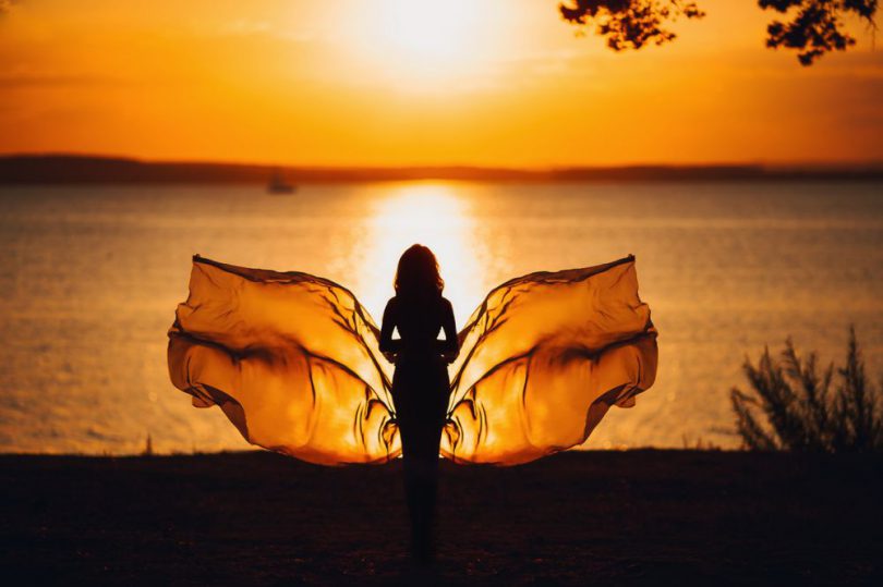 Mulher com "asas" de borboleta em frente ao mar no pôr-do-sol.