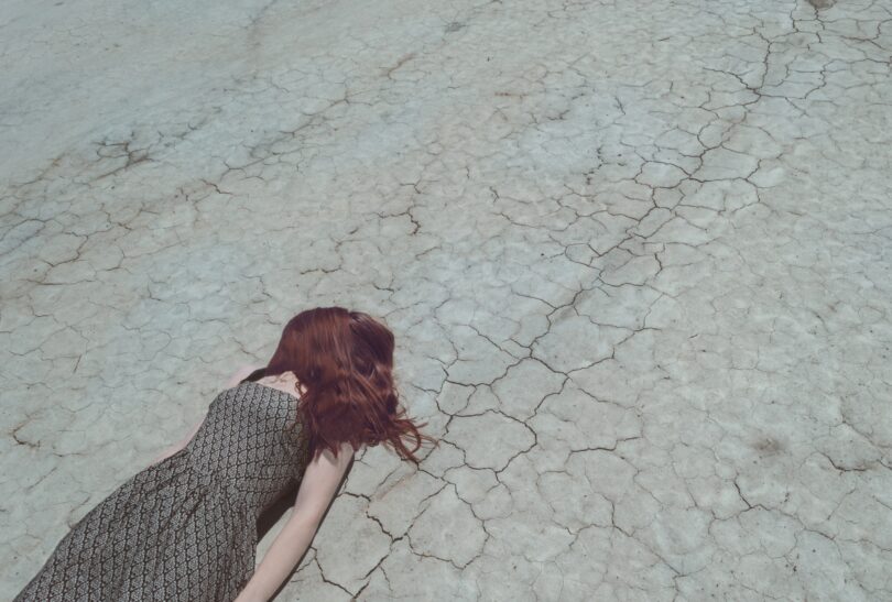 Garota da pele branca e cabelos ruivos vestindo um vestido estampado com detalhes em marrom e branco. Ela está deitada sobre um chão rachado com seus cabelos cobrindo o rosto.