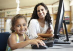 Professora e aluna sorrindo sentadas de frente a um computador.