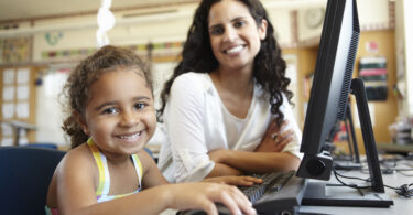 Professora e aluna sorrindo sentadas de frente a um computador.