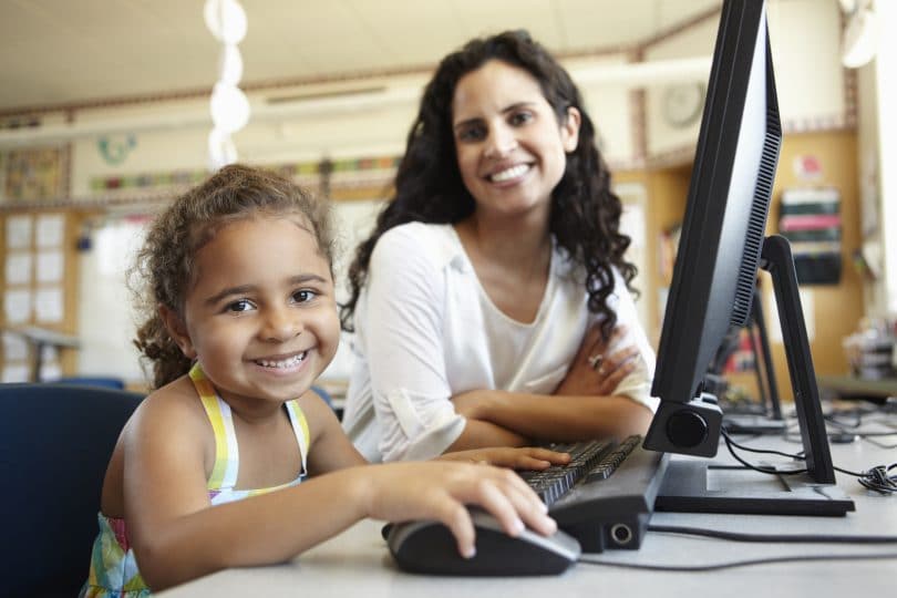 Professora e aluna sorrindo sentadas de frente a um computador.