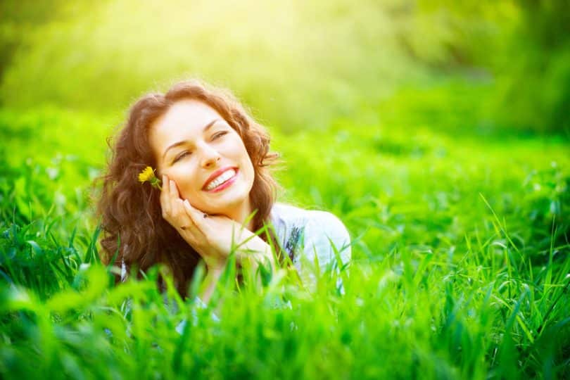 Mulher na grama com mãos o rosto sorrindo e sol refletindo