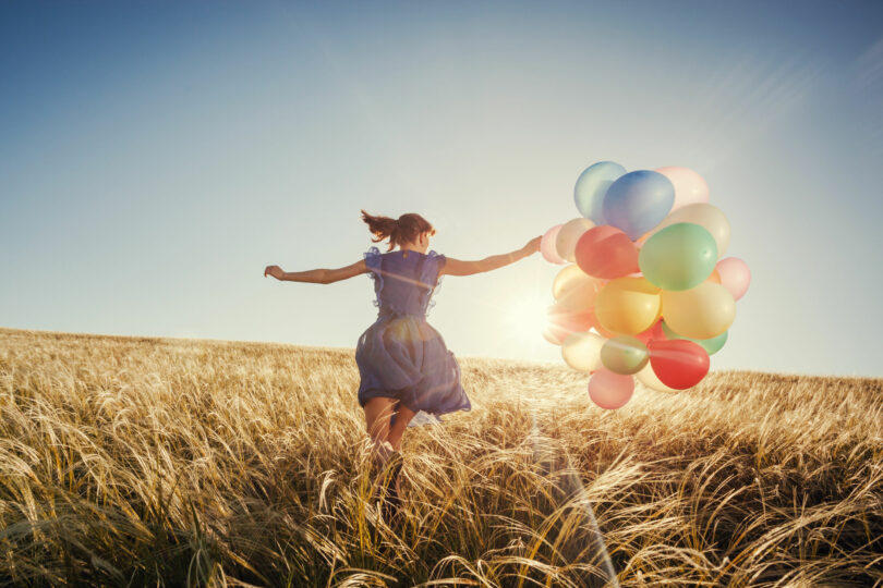 Menina correndo no campo com balões ao pôr do sol.