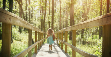 Menina correndo em ponte de madeira no meio de floresta durante o dia.