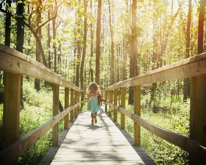 Menina correndo em ponte de madeira no meio de floresta durante o dia.