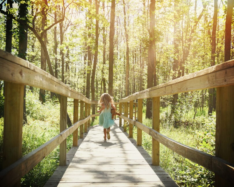 Menina correndo em ponte de madeira no meio de floresta durante o dia.