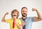 Pai e filho, ambos e brancos, segurando um bigode feito de papel em frente do rosto, dobrando o braço em sinal de força.