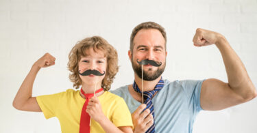 Pai e filho, ambos e brancos, segurando um bigode feito de papel em frente do rosto, dobrando o braço em sinal de força.