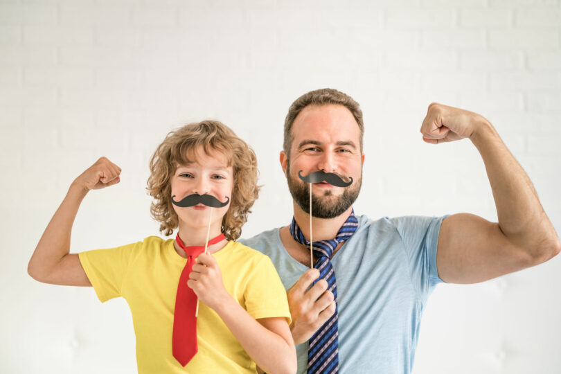 Pai e filho, ambos e brancos, segurando um bigode feito de papel em frente do rosto, dobrando o braço em sinal de força.