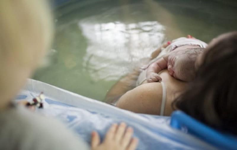 Bebê recém-nascido deitado no colo da mãe que está na banheira.