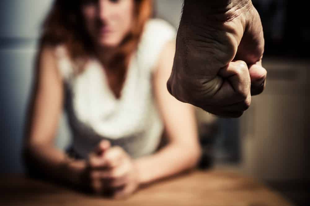 Mulher sentada em uma mesa segurando as próprias mãos olhando a mão de um homem cerrada representando a violência doméstica