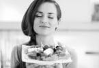 Foto preta e branca de mulher sorridente, de olhos fechados, segurando um prato de salada.