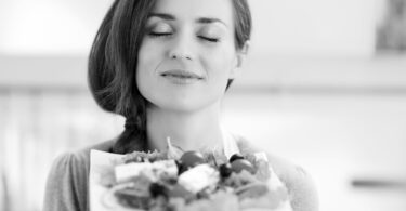 Foto preta e branca de mulher sorridente, de olhos fechados, segurando um prato de salada.