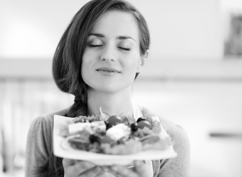 Foto preta e branca de mulher sorridente, de olhos fechados, segurando um prato de salada.