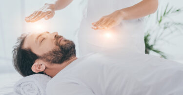 Homem branco, jovem, barbudo, vestido de branco, deitado em uma maca, com uma outra pessoa em pé ao seu lado, colocando as mãos sobre ele, em uma sessão de reiki.