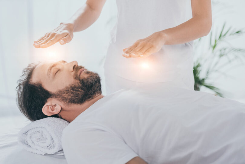 Homem branco, jovem, barbudo, vestido de branco, deitado em uma maca, com uma outra pessoa em pé ao seu lado, colocando as mãos sobre ele, em uma sessão de reiki.