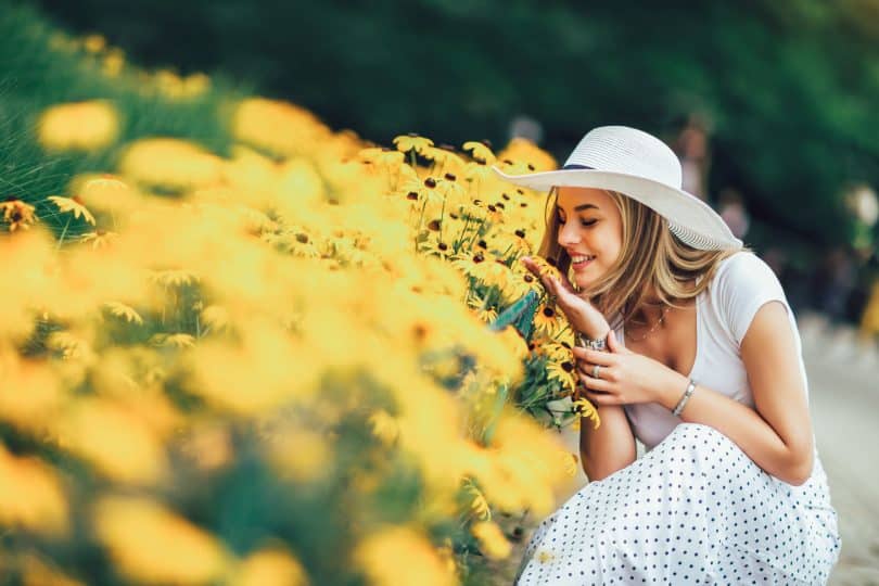 Mulher jovem sorrindo ao lado de um canteiro de flores amarelas.