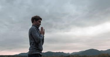 Homem jovem, em pé perto de montanhas, com as mãos cruzadas em sinal de oração, representando o despertar da essência.