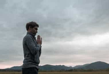 Homem jovem, em pé perto de montanhas, com as mãos cruzadas em sinal de oração, representando o despertar da essência.