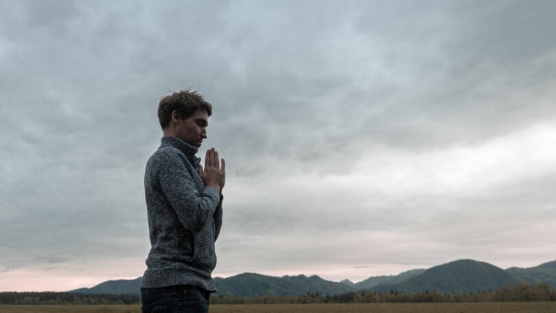 Homem jovem, em pé perto de montanhas, com as mãos cruzadas em sinal de oração, representando o despertar da essência.