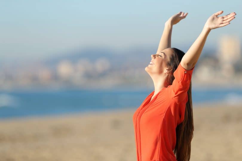 Foto de mulher feliz, jovem, com os braços para cima, em uma praia. Representando liberdade e autonomia.