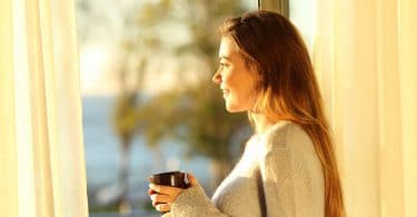 Mulher olhando pela janela com sol refletindo segurando xícara de café