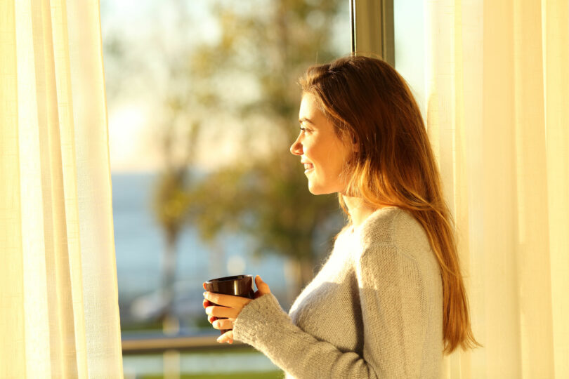 Mulher olhando pela janela com sol refletindo segurando xícara de café