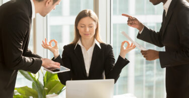 Mulher meditando em sua mesa de trabalho enquanto dois homens ao gritam ao redor dela.