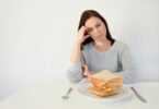 Mulher jovem, branca, triste, vestindo um moletom cinza, sentada em uma mesa, olhando para um prato cheio de fatias de pães.