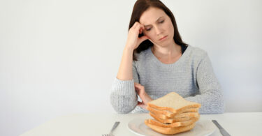 Mulher jovem, branca, triste, vestindo um moletom cinza, sentada em uma mesa, olhando para um prato cheio de fatias de pães.