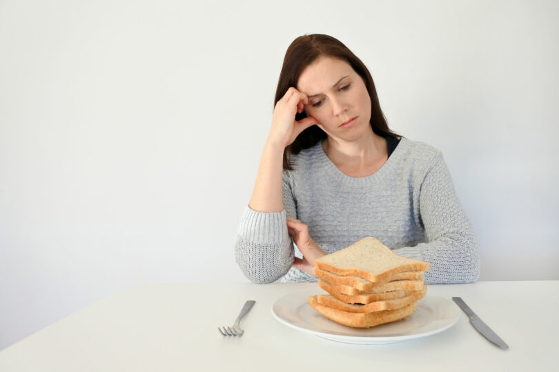 Mulher jovem, branca, triste, vestindo um moletom cinza, sentada em uma mesa, olhando para um prato cheio de fatias de pães.