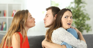 Homem jovem branco, abraçado à sua namorada, também branca e jovem, e, ao mesmo tempo, tentando beijar outra menina, também branca e jovem. Todas as pessoas estão sentadas no sofá de uma sala.