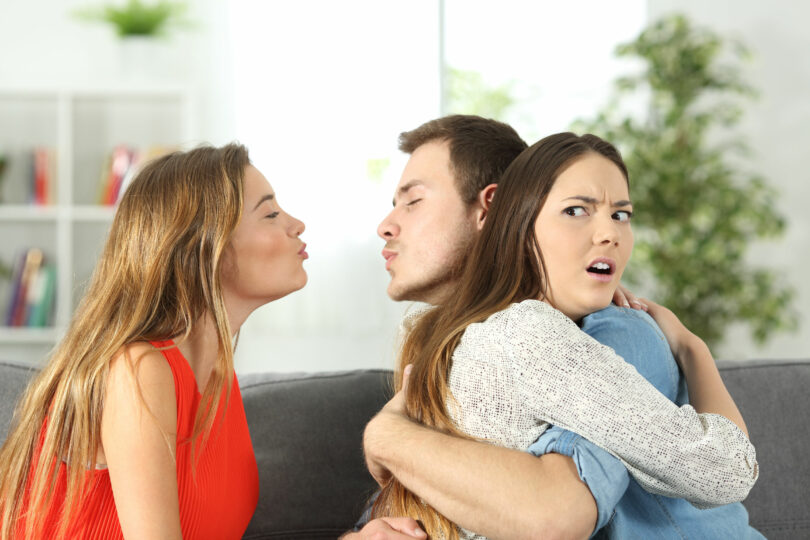 Homem jovem branco, abraçado à sua namorada, também branca e jovem, e, ao mesmo tempo, tentando beijar outra menina, também branca e jovem. Todas as pessoas estão sentadas no sofá de uma sala.