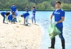 Grupo de voluntários, todos vestindo camisetas azuis, recolhendo lixo da areia da praia, segurando sacos de lixo.