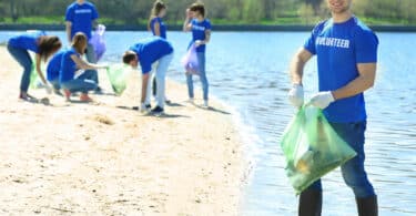 Grupo de voluntários, todos vestindo camisetas azuis, recolhendo lixo da areia da praia, segurando sacos de lixo.