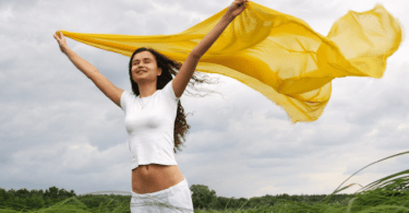 Mulher segurando um véu amarelo enquanto sente a brisa em um jardim.