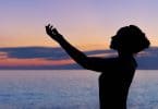 Silhueta de mulher com as mãos para cima, buscando cura, em uma paisagem de pôr do sol no mar.