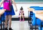 Menina em pé na sala de aula fazendo bullying com sua colega de classe, que chora no chão, sentada.